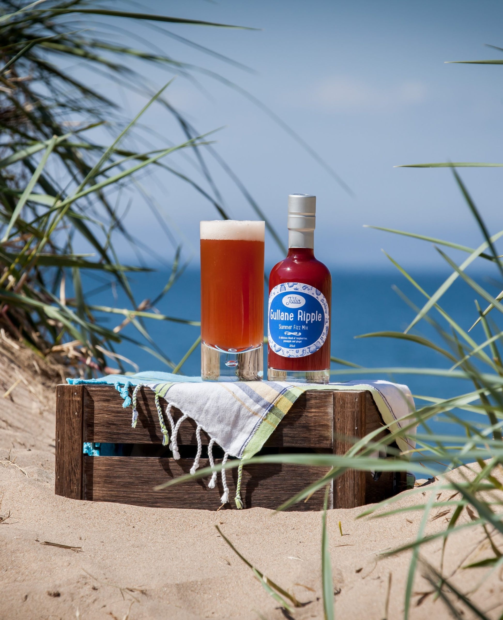 Large Bottle of Gullane Ripple Summer Fizz Mix and glass of cocktail on wooden crate with beach in background