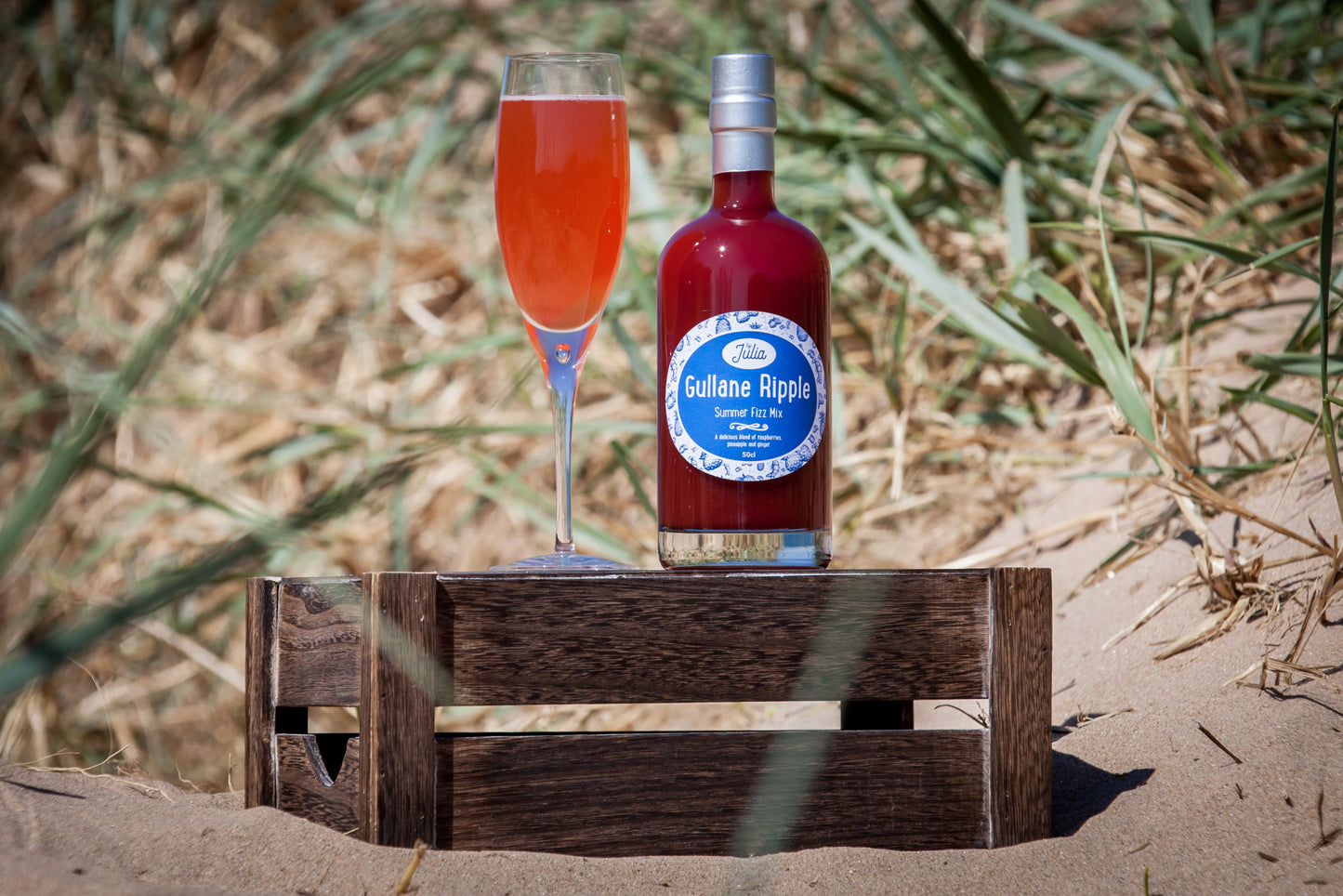 Large Bottle of Gullane Ripple Summer Fizz Mix and glass of cocktail on wooden crate with beach in background