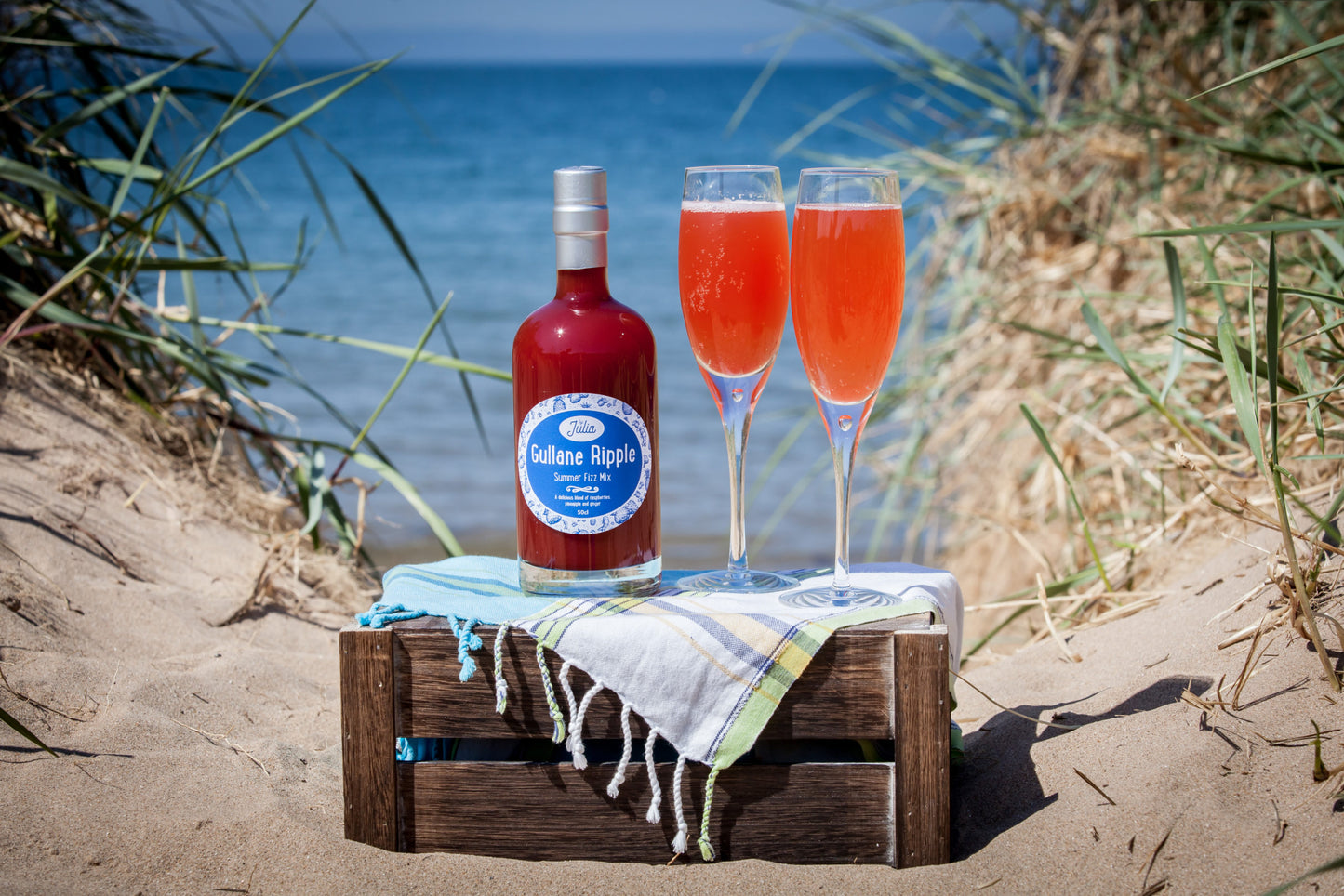 Large Bottle of Gullane Ripple Summer Fizz Mix and two glasses of cocktail on wooden crate with beach in background