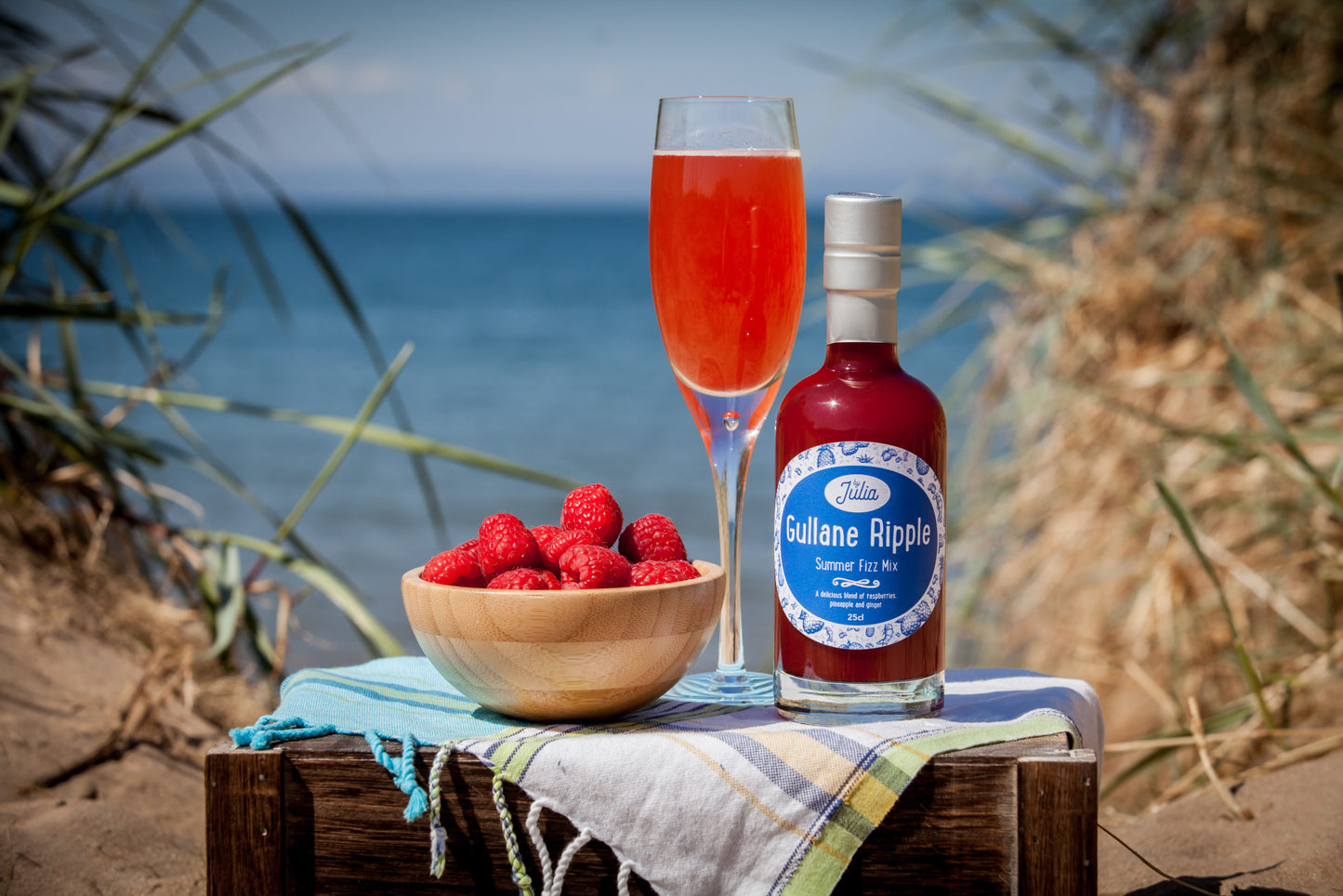 Large Bottle of Gullane Ripple Summer Fizz Mix and glass of cocktail on wooden crate with beach in background and bowl of raspberries